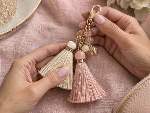 Illustrative AI image: hands holding blush and cream tassel charms with a gold chain and clasp