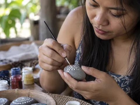 Illustrative AI image: hands painting white dots onto a smooth grey stone with a tiny brush